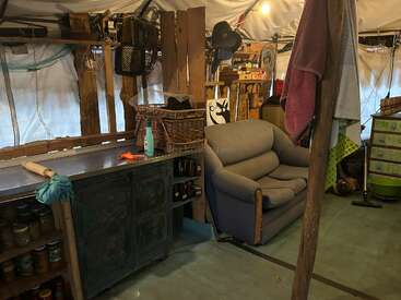 Une pièce chaleureuse et rustique, avec des armoires en bois, des étagères remplies de bocaux, un fauteuil confortable, des serviettes suspendues, des paniers et divers objets du quotidien, le tout disposé dans un espace qui respire la vie de tous les jours.