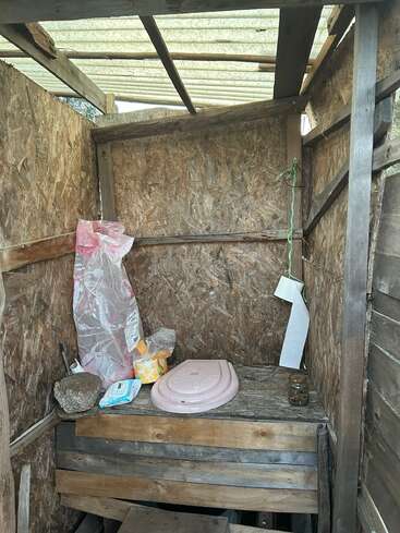 Cette image montre des toilettes extérieures rustiques en bois, dotées d'un toit, d'un siège de toilettes, de produits d'entretien, d'un rouleau de papier toilette suspendu et de divers objets posés sur le banc en bois.