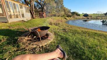A person relaxes by a fire pit near a pond, with a rustic cabin, trees, and green grass under bright sunshine in a peaceful countryside setting.