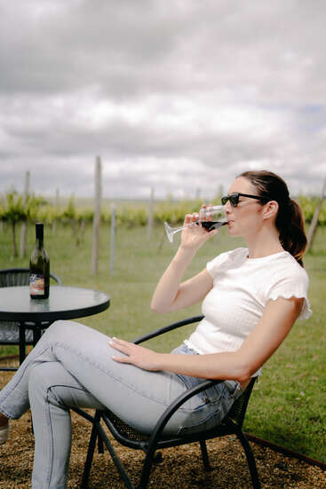 A woman wearing sunglasses relaxes on a patio chair at a vineyard, enjoying a glass of red wine with a wine bottle on the table beside her.