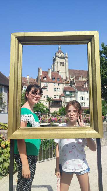 Zwei Mädchen posieren glücklich lächelnd in einem großen goldenen Rahmen vor einem Hintergrund aus charmanten europäischen Gebäuden und einem hohen Turm unter einem klaren blauen Himmel.