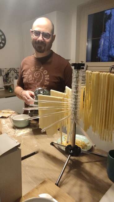 Un homme portant des lunettes et un maillot marron du FC Barcelone sourit dans une cuisine, en train de préparer des pâtes maison à l'aide d'une machine à pâtes et d'un séchoir. Il fait nuit dehors.