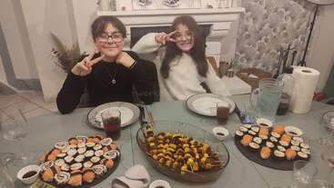 Deux enfants souriants posent en faisant le signe de la paix devant une table dressée avec des sushis, des brochettes, des boissons et des assiettes, créant une atmosphère chaleureuse et joyeuse.