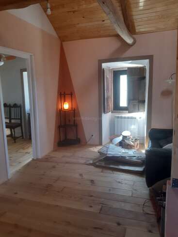 A cozy room with wooden floors and ceiling, exposed beam, armchair, window with shutters, Himalayan lamp on shelf, scattered boxes, and adjacent rooms visible.