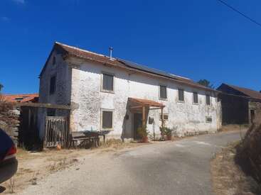 A rustic, old white house with a tiled roof, solar panels, and multiple windows. There are plants, a small bench, and bright blue sky overhead.