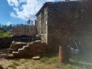 Das Bild zeigt ein altes Steinhaus mit verwitterten Wänden, ein rostiges Fass, Gartenstühle, eine Schubkarre, grobe Steinstufen und einen strahlend blauen Himmel.