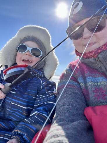 The image shows a woman and a child in winter clothing, with the child holding a fishing rod and reel, set against a sunny blue sky.