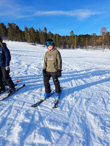 The image depicts a young boy standing on skis, wearing a helmet and goggles, with a woman in the background, set against a snowy landscape with trees and a blue sky.