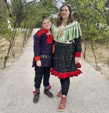 The image depicts a woman and a boy standing on a path, both dressed in traditional clothing, with the woman wearing a green shawl and the boy sporting a dark blue shirt with red accents.