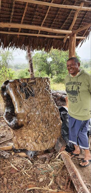 Ein lächelnder Mann steht neben einem großen, kunstvoll geschnitzten Baumstamm unter einem strohgedeckten Unterstand, umgeben von der Natur und mit beeindruckender Handwerkskunst und künstlerischem Talent.