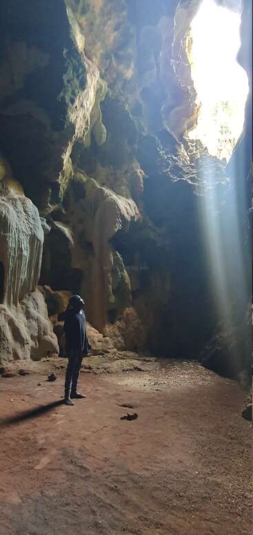 Eine Person steht in einer großen, schwach beleuchteten Höhle. Das Sonnenlicht, das durch eine Öffnung von oben einfällt, beleuchtet die Felswände und schafft eine dramatische, ehrfurchtgebietende Szene.