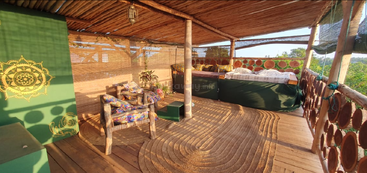 Dieser gemütliche, rustikale Außenraum hat Holzmöbel, ein bequemes Bett, bunte Kissen, einen Teppich aus Naturfasern, eine Bambusdecke, üppiges Grün und viel Tageslicht.