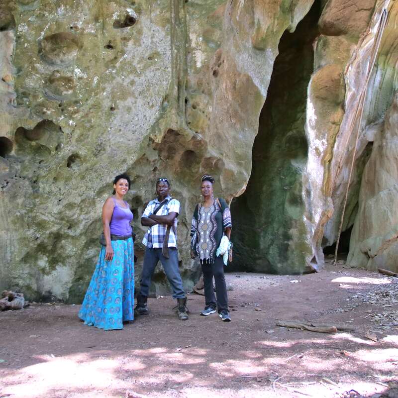 Drei Menschen stehen zusammen vor einer großen, strukturierten Felsformation mit Höhlen. Sie wirken entspannt und glücklich und tragen bequeme Outdoor-Kleidung.