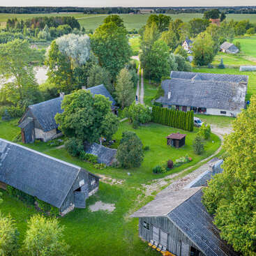 Das Bild zeigt eine ruhige, ländliche Landschaft mit einer Ansammlung rustikaler Gebäude, üppigem Grün und einem gewundenen Weg vor einer malerischen Kulisse aus Bäumen und Feldern.