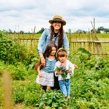 Una mujer y dos niños permanecen juntos en un exuberante jardín, sonriendo y disfrutando de la naturaleza. Los niños sostienen flores, rodeados de plantas verdes y vallas rústicas.