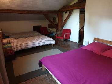 Cette chambre mansardée confortable comprend des poutres en bois apparentes, deux lits avec des literies différentes, deux chaises rouges, un tapis, des planchers en bois et un plafond partiellement incliné.