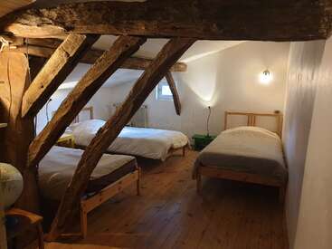 Cette chambre mansardée confortable dispose de trois lits simples, de poutres en bois rustiques, d'un plancher en bois, de murs blancs, d'une petite fenêtre et d'un mobilier simple, créant ainsi une atmosphère chaleureuse et accueillante.