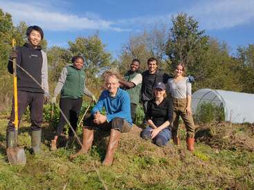 Un groupe diversifié de sept personnes pose en plein air dans une ferme, souriant, portant des bottes et des gants, entouré de verdure, avec une serre visible à l'arrière-plan.