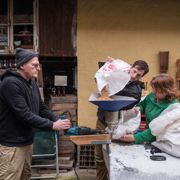 L'image représente trois personnes réunies autour d'une machine, l'une d'elles y versant un sac de céréales, sur fond d'atelier rustique en plein air.