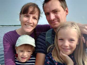 Una feliz familia de cuatro miembros posa para una foto de primer plano al aire libre, junto a un lago. Los padres sonríen con cariño, junto a sus dos hijos pequeños, disfrutando juntos del momento.