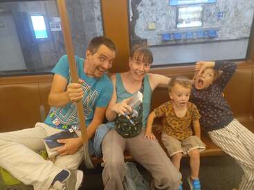 Una familia de cuatro miembros está sentada en un banco del metro haciendo muecas y gestos graciosos. Parecen felices y juguetones, disfrutando juntos del trayecto. Al fondo se ve una estación.