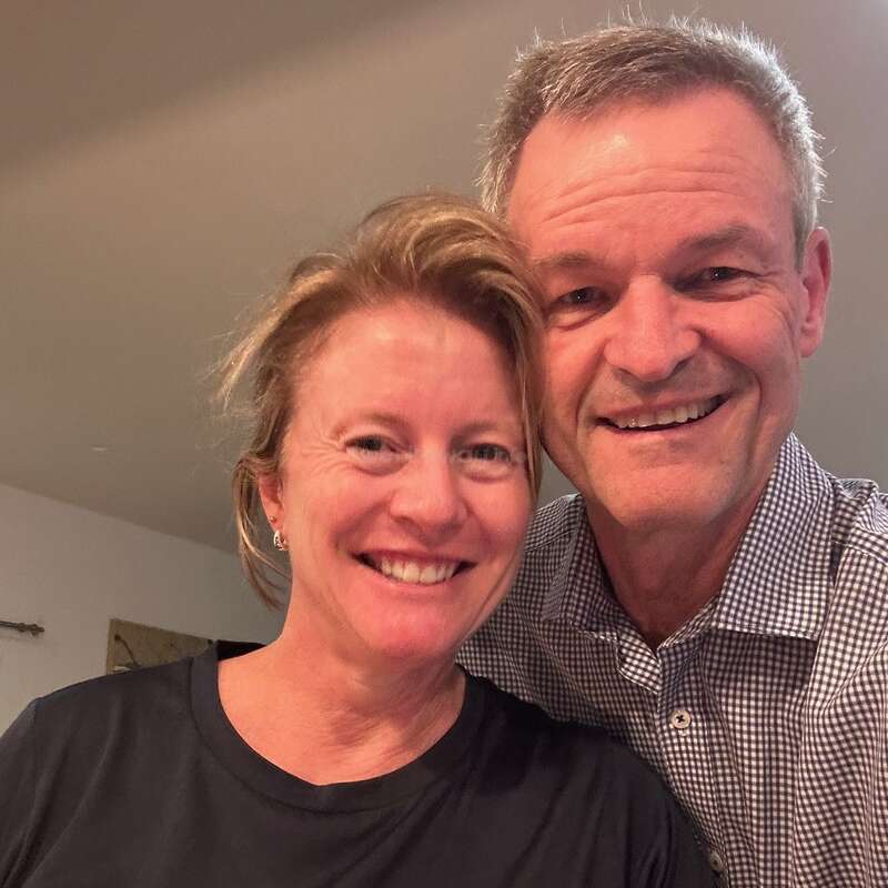 Das Bild zeigt ein Paar mittleren Alters, das für ein Selfie posiert. Die Frau auf der linken Seite trägt ein schwarzes Fila-T-Shirt und der Mann auf der rechten Seite ein kariertes Hemd.