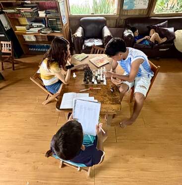 Drei Kinder sitzen um einen Holztisch, spielen Schach und schreiben in Hefte. Um sie herum stehen Bücherregale und Sofas. Ein anderes Kind entspannt sich auf dem Sofa und liest.