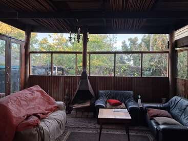 A cozy porch with large windows, rustic wooden decor, leather sofas, a central fireplace, coffee table, and tree views, blending indoor comfort with nature outside.