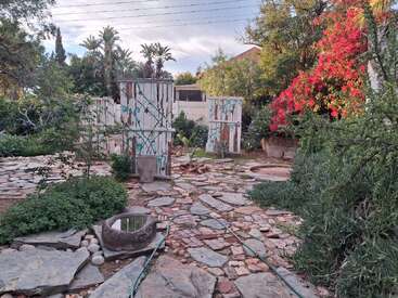 A rustic garden with stone pathways, lush greenery, blooming pink flowers, scattered plants, painted wooden structures, and a hose laying across vibrant, natural landscaping. Peaceful ambiance.