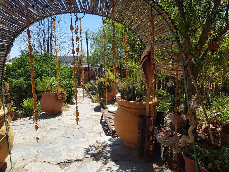 The image depicts a serene outdoor setting, featuring a stone pathway, potted plants, and a wooden arched structure adorned with hanging orange decorations, set against a clear blue sky.