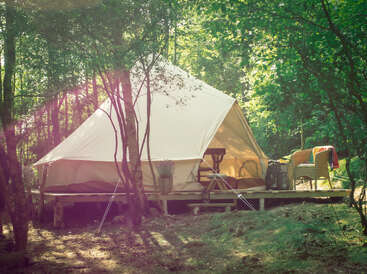 Uma aconchegante barraca de glamping está instalada em uma plataforma de madeira em uma floresta iluminada pelo sol. Cadeiras confortáveis e luz solar suave criam um retiro tranquilo, convidativo e natural.