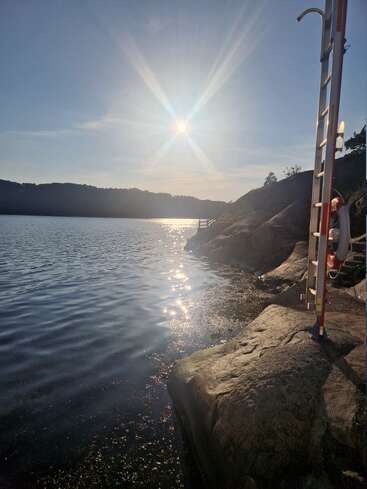 La imagen capta una serena escena junto a un lago al atardecer, con la luz del sol reflejándose en aguas tranquilas, la orilla rocosa, una escalera y un ambiente tranquilo y natural.