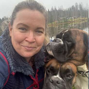 A woman smiles while taking a selfie outdoors with two brown and white boxers. In the background, there is a lake and some trees.