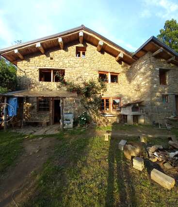 Das Bild zeigt ein rustikales Steinhaus mit einem Giebeldach, mehreren Fenstern und einer überdachten Veranda inmitten einer ruhigen natürlichen Umgebung.