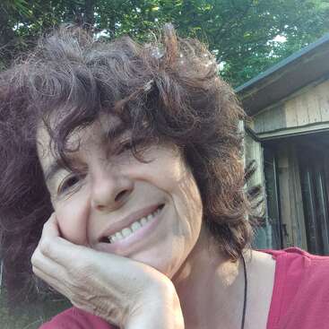 Das Bild zeigt eine Frau mit lockigen braunen Haaren, die ein rosa Hemd trägt und sich auf ihre Hand stützt, vor dem Hintergrund eines Gebäudes und von Bäumen.