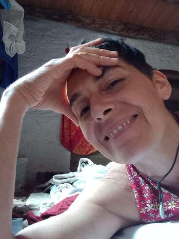 Das Bild zeigt eine Frau mit kurzen dunklen Haaren, die ein rotes geblümtes Oberteil und eine Halskette trägt und ihre Hand in einer entspannten Pose auf die Stirn legt.