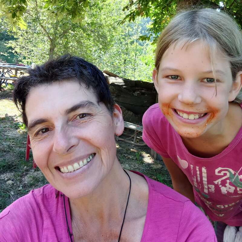 Das Bild zeigt eine Frau und ein Mädchen, die für ein Selfie in einem Waldgebiet posieren, wobei die Frau auf der linken und das Mädchen auf der rechten Seite steht.