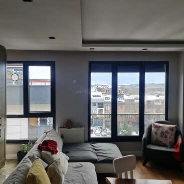 A cozy living room with a sectional sofa, armchair, and pillows. Large windows offer a view of nearby buildings and snow-covered rooftops, creating a serene atmosphere.