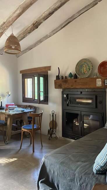 Esta acogedora habitación rústica cuenta con vigas de madera a la vista, una estufa de leña, una pequeña mesa de comedor, sillas sencillas, platos decorativos, una ventana y una iluminación suave.