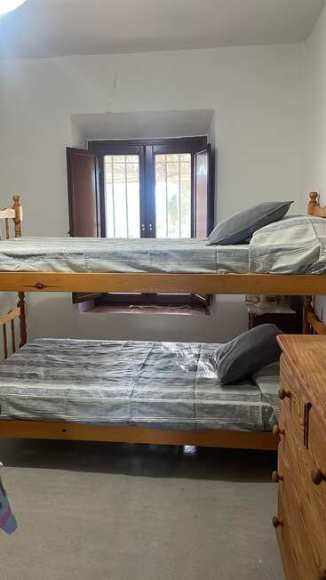 Esta acogedora habitación cuenta con literas de madera con ropa de cama a rayas grises, una pequeña cómoda de madera, una ventana abierta con contraventanas y luz que entra a raudales, creando un ambiente tranquilo.