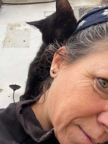 Un juguetón gato negro se sube al hombro de una mujer y le acaricia el pelo con el hocico. La mujer, que lleva una diadema azul, sonríe levemente en la selfie tomada en primer plano.