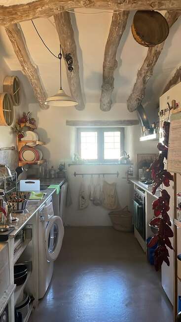 Eine gemütliche, rustikale Küche mit sichtbaren Holzbalken, Tageslicht durch ein Fenster, hängenden Kräutern, Regalen mit Geschirr und einer Waschmaschine unter der Arbeitsplatte.