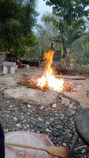 Ein Lagerfeuer brennt hell in einer rustikalen Umgebung im Freien, umgeben von Steinen, Pflanzen und Bänken. Im Vordergrund sind ein Holzlöffel und ein Behälter zu sehen.