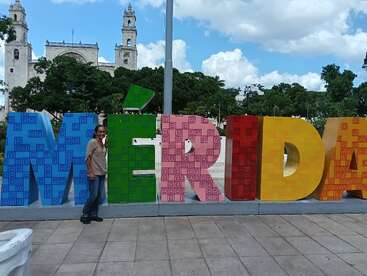 Eine Person steht lächelnd neben großen bunten "MÉRIDA"-Buchstaben auf einem Platz. Dahinter zieren Bäume, eine historische Kirche und ein strahlend blauer Himmel die Szene.