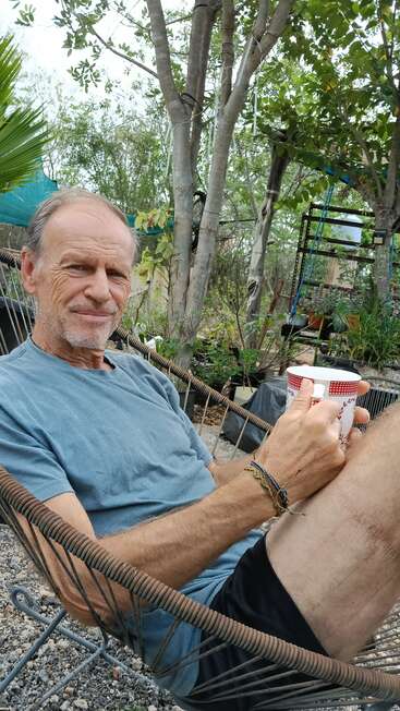 Ein entspannter älterer Mann sitzt bequem in einer Hängematte im Freien und hält einen gemusterten Becher in der Hand. Um ihn herum sind Bäume, Grünpflanzen und eine gartenähnliche, ruhige Atmosphäre.