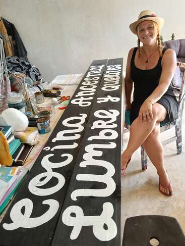 Eine lächelnde Frau sitzt neben einem handgemalten Holzschild mit der Aufschrift "Cocina Ancestral Lumbre y Querencias". Auf dem Tisch liegen Farben, Pinsel und andere kreative Utensilien.