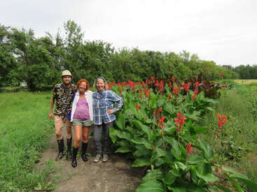 Three people stand together outdoors, smiling near a lush garden bed filled with tall green plants and vibrant red flowers. Trees and greenery fill the background.