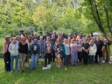 A large group of people, men and women of various ages, stand outdoors on green grass, smiling for a group photo with trees in the background.
