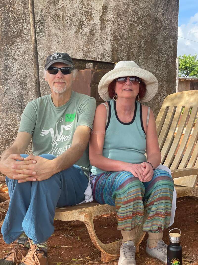 A couple relaxes on a rustic wooden bench outdoors. Both wear sunglasses and hats. They are dressed casually and colorfully, enjoying a sunny, peaceful moment together.