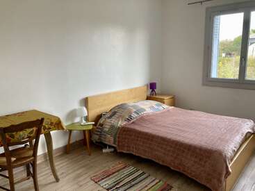 A cozy bedroom with a wooden bed, colorful bedding, small bedside tables, a window, a rug, a table, and a chair in natural light.